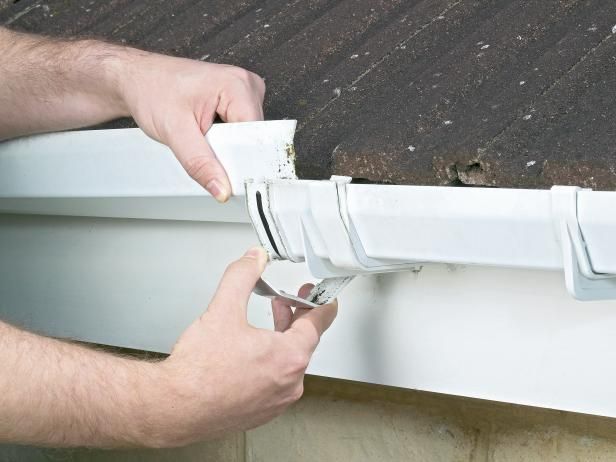 Hands fitting a white gutter downspout section with a clamp on a house exterior.