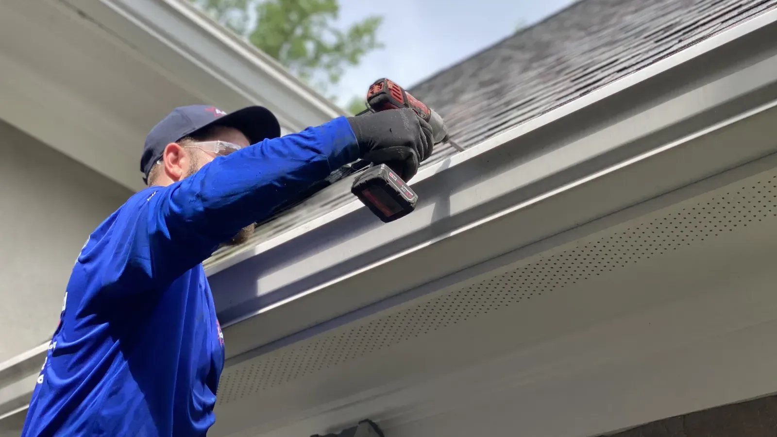 Worker in blue uniform using a power drill on a house gutter under the eaves