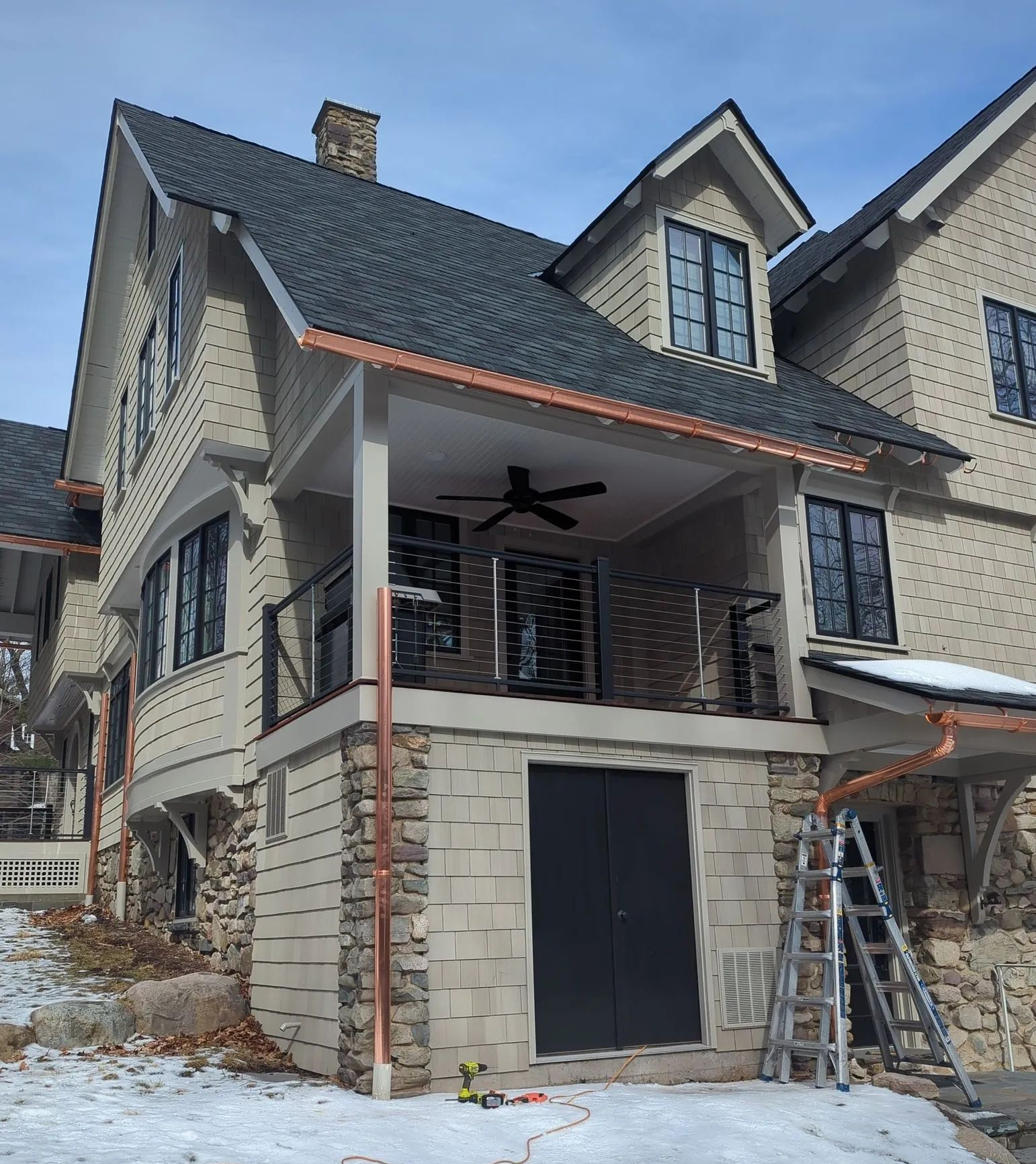 Two-story stone house with a covered porch, dark roof, and a ladder in the snowy yard