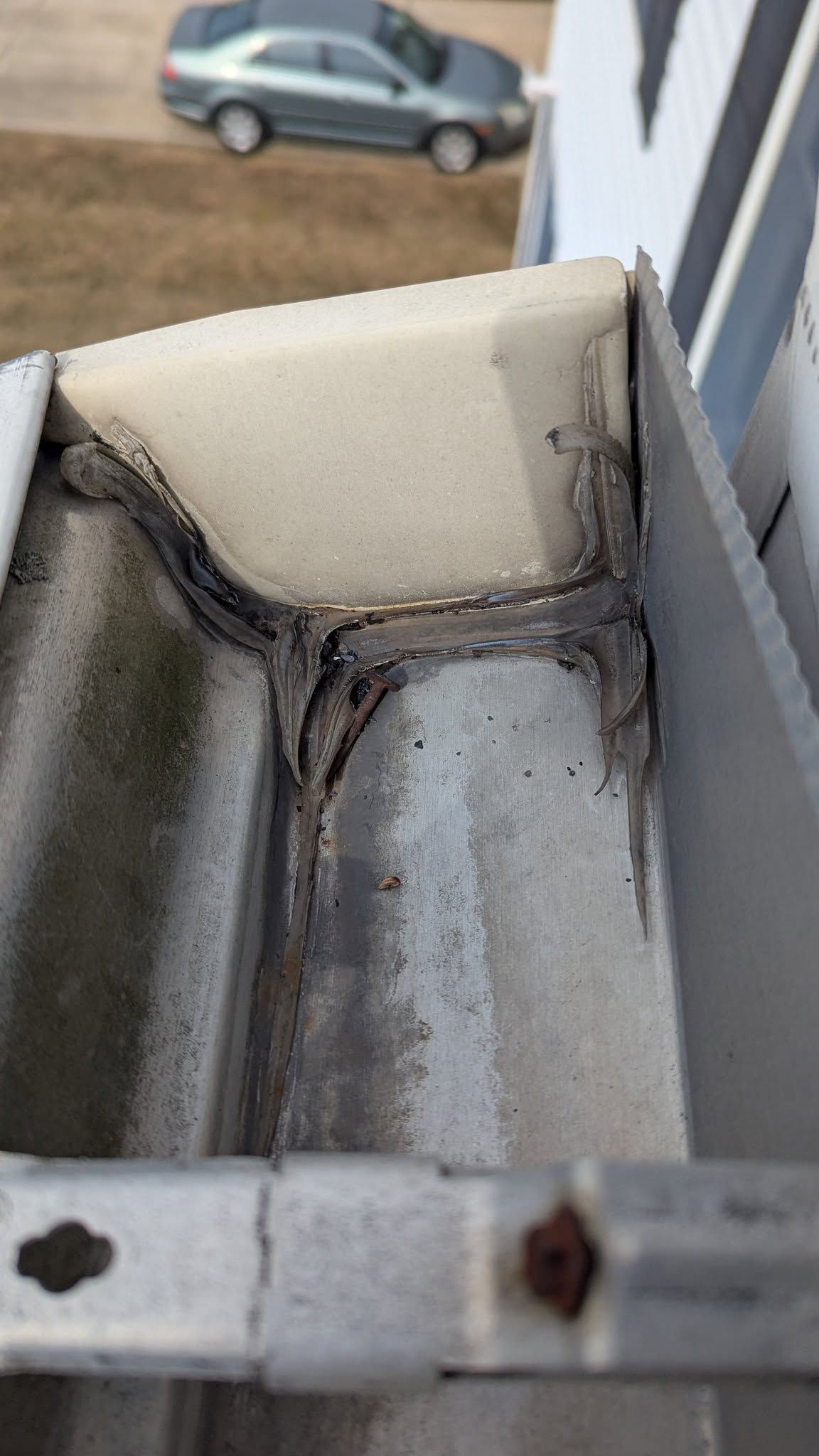 Damaged gutter corner with peeling sealant and rust, viewed from a rooftop with a car below.