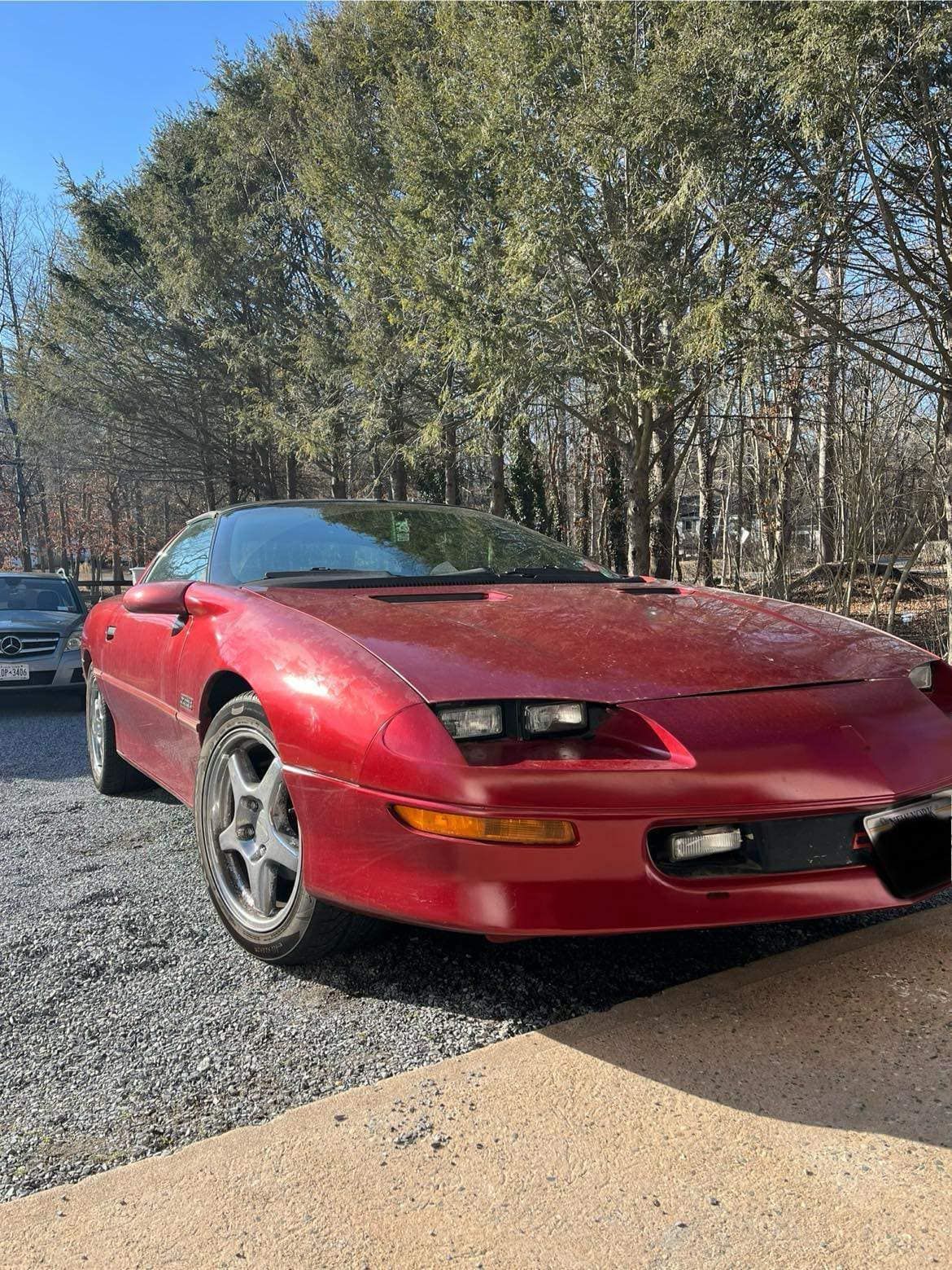 A red car is parked in a gravel lot in front of trees.