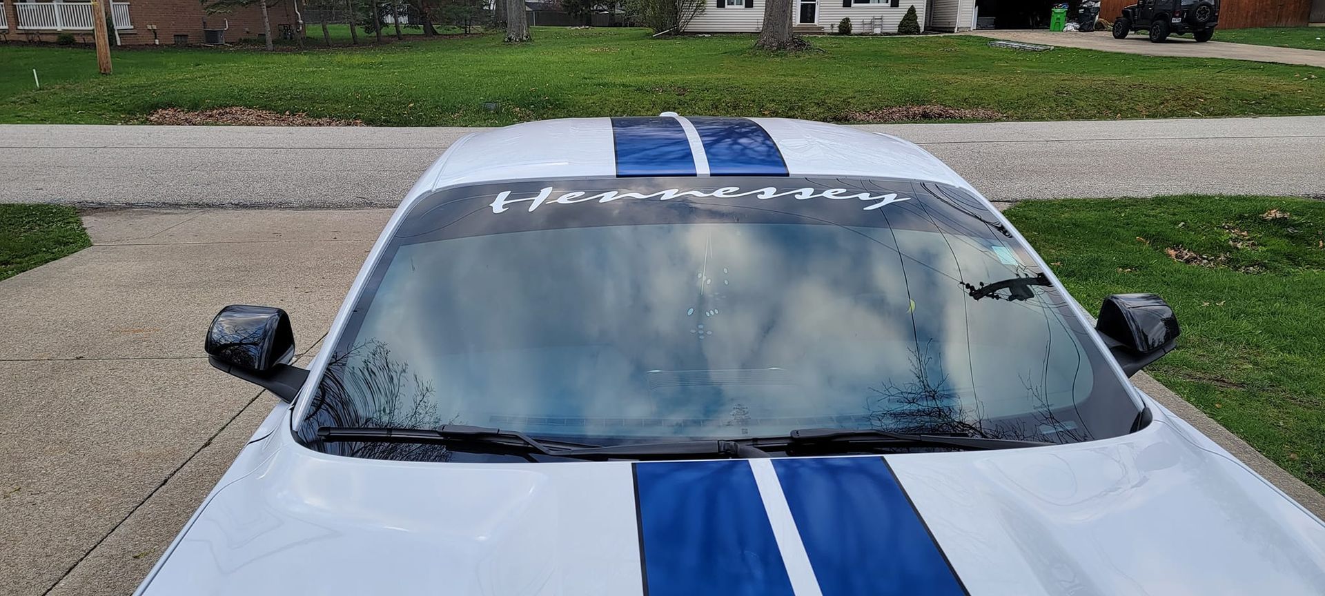 A white mustang with blue stripes on the hood is parked in a driveway.