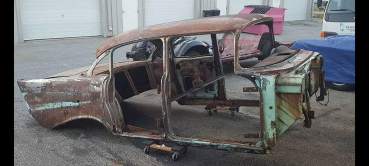 A rusty old car is sitting on the ground in front of a garage.