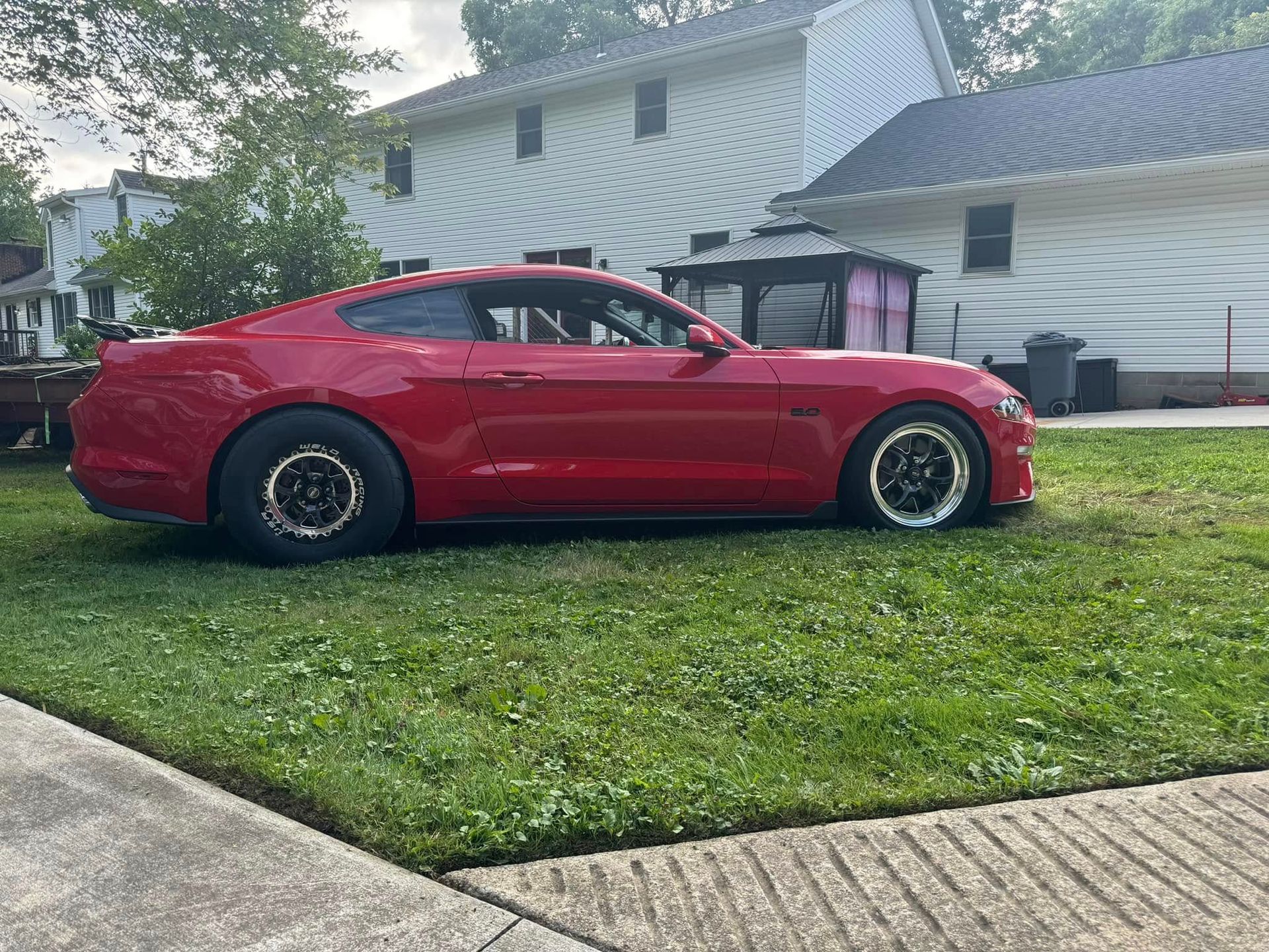 A red mustang is parked in the grass in front of a house.