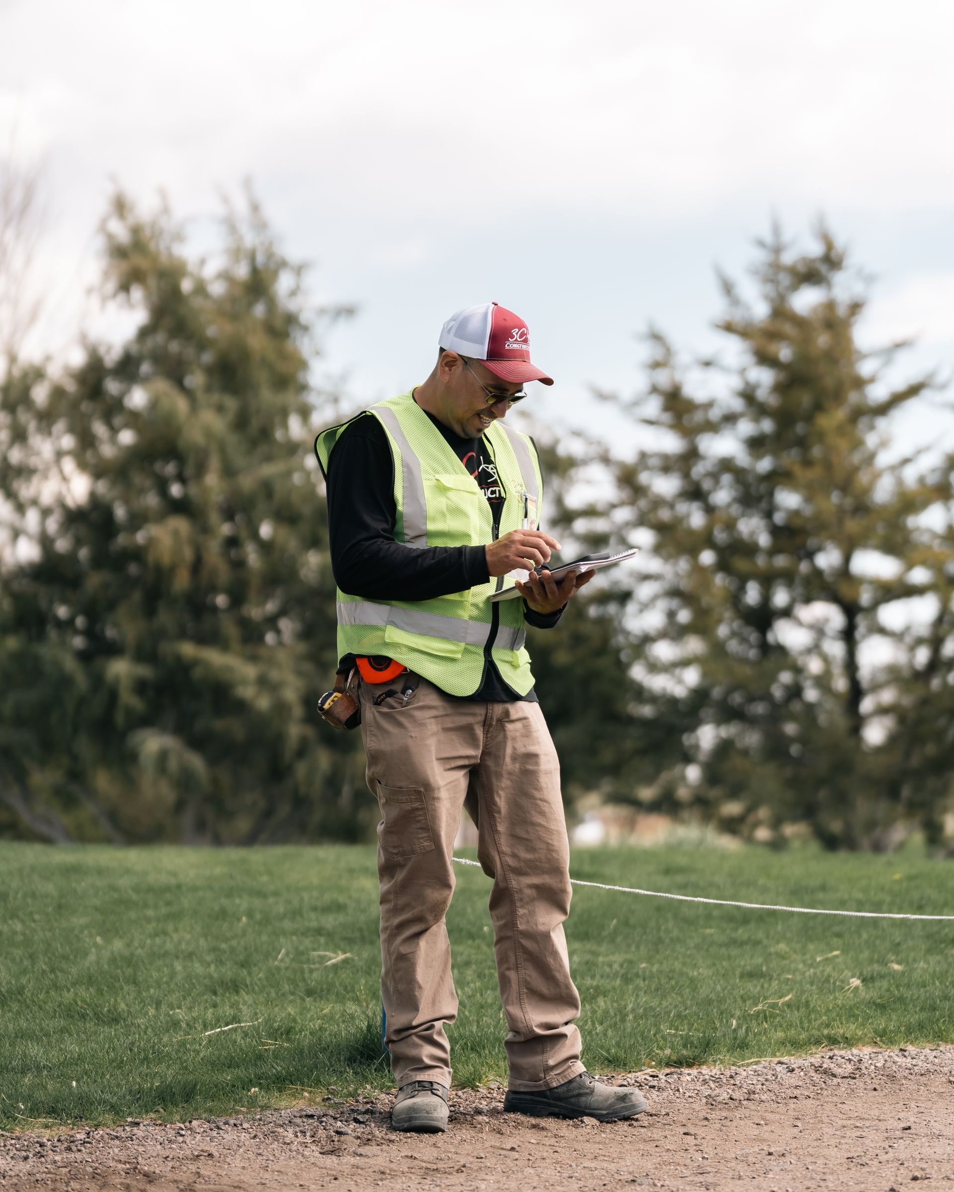 Man in safety vest, hat, and work pants, taking notes outdoors on a cloudy day.