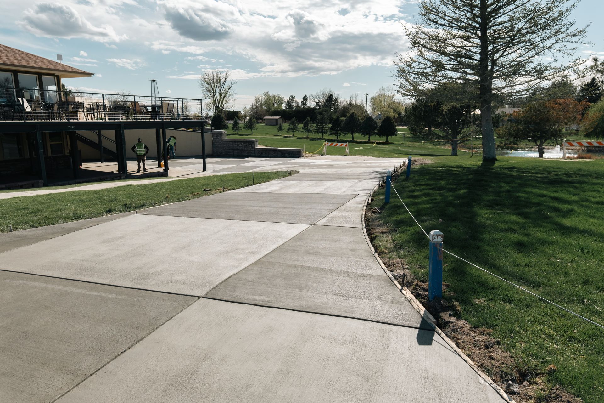 Concrete pathway with blue posts and rope along grassy area, near a building and trees under a cloudy sky.