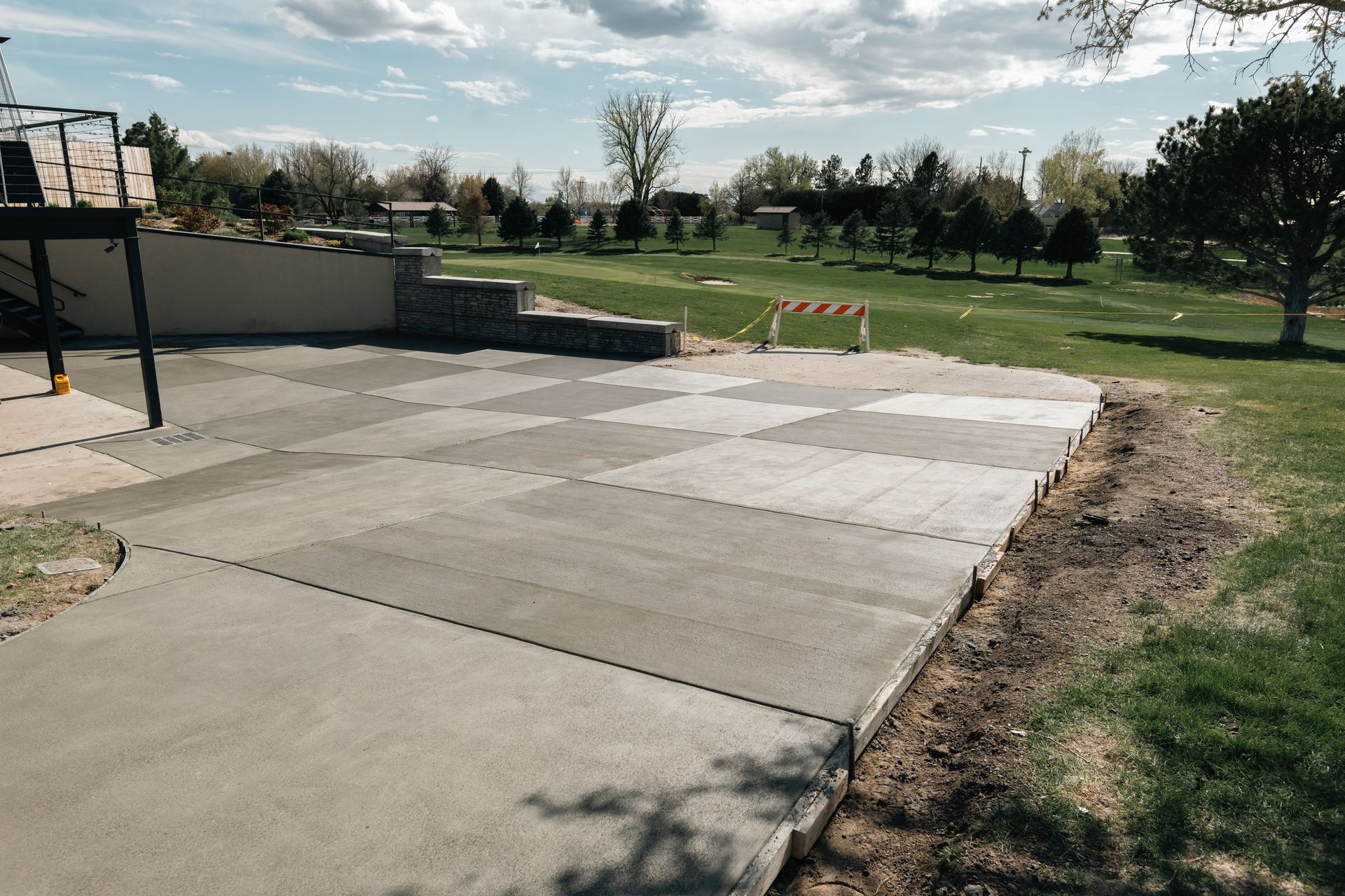 Concrete patio with checkered tile section next to a grassy area, small trees, and a retaining wall.