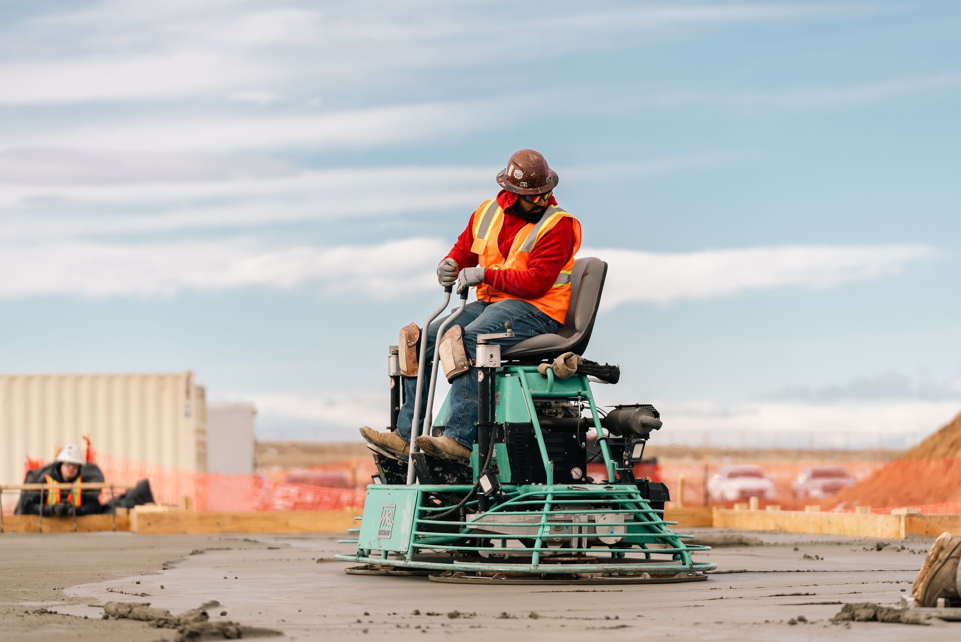 Worker in safety vest operating a ride-on concrete finishing machine on a construction site.