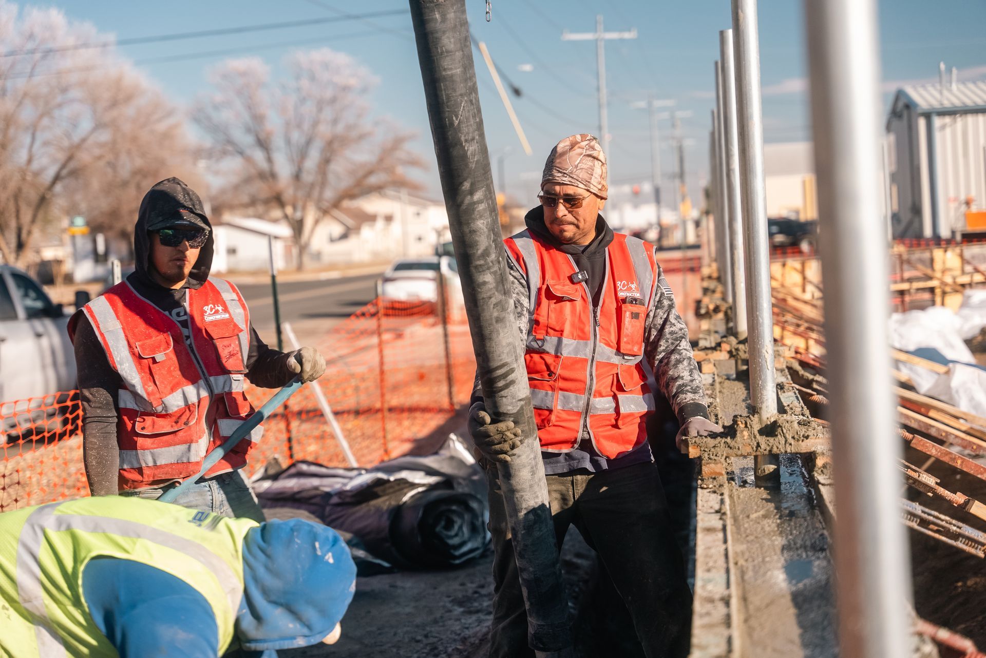 Construction workers pouring concrete on a sunny day. Two wear orange vests, one directs the hose.