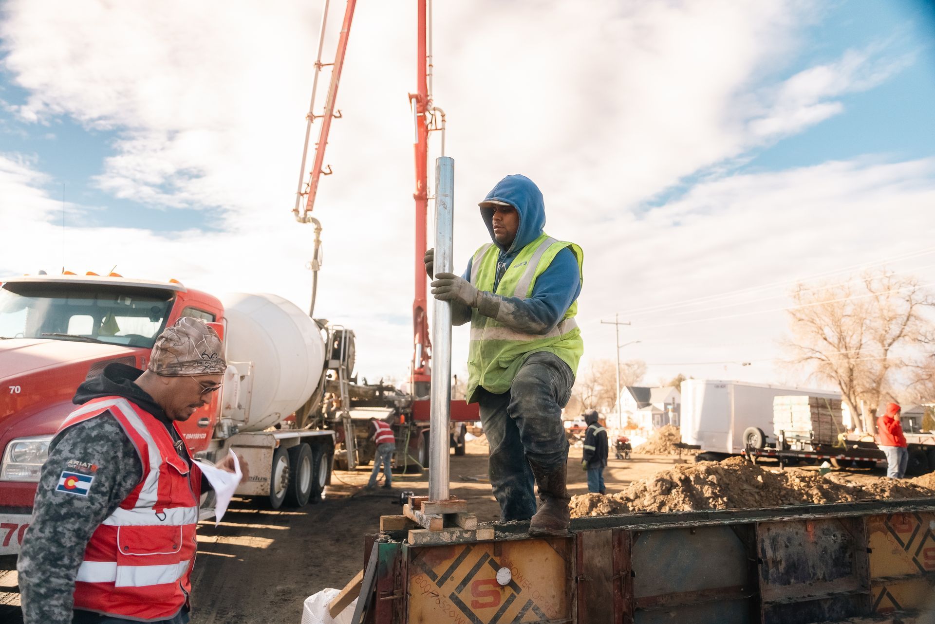 Two construction workers at a concrete pour, one holding a metal rod. A red cement truck and pump are visible.