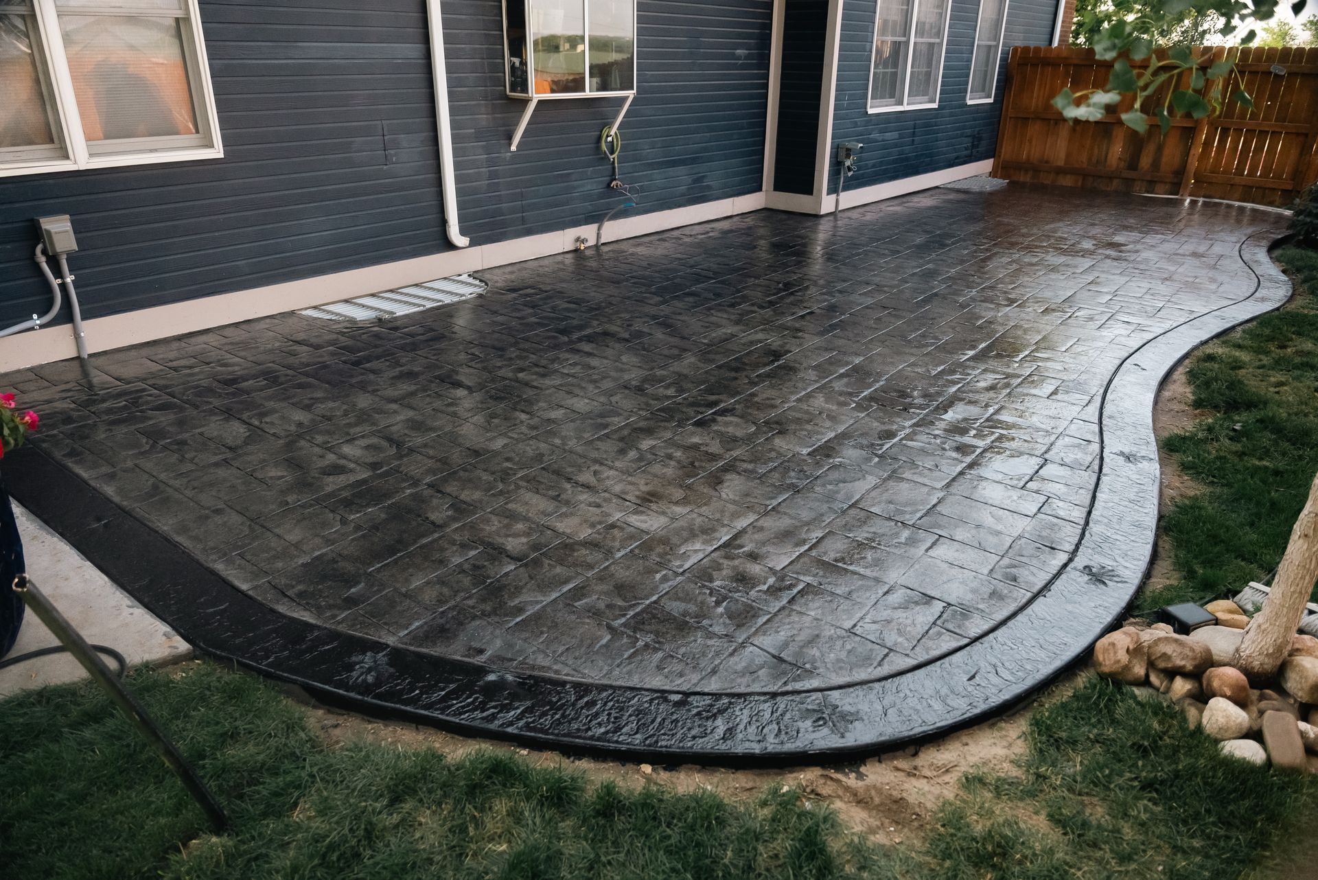 Dark gray stamped concrete patio with curved border next to a dark blue house and lawn.
