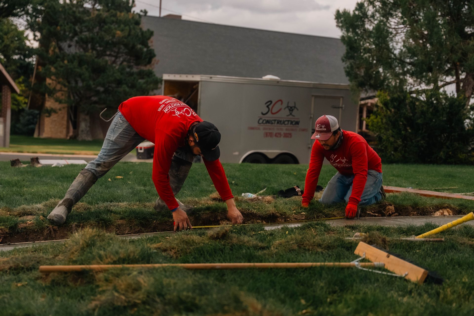 Two construction workers in red shirts work on a lawn. One bends over, the other kneels. A trailer sits in the background.