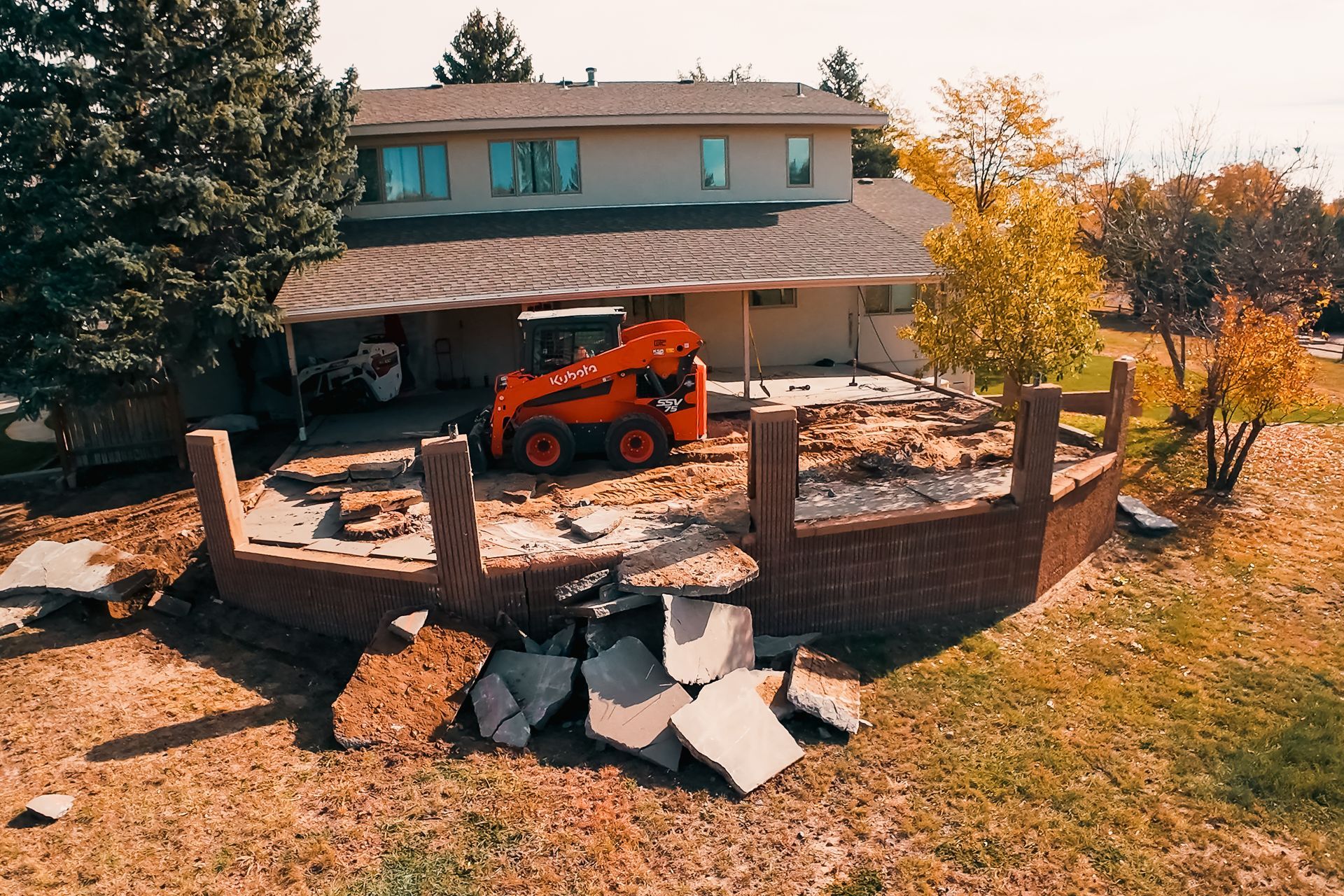 A small orange skid steer demolishing a brick patio near a two-story house.