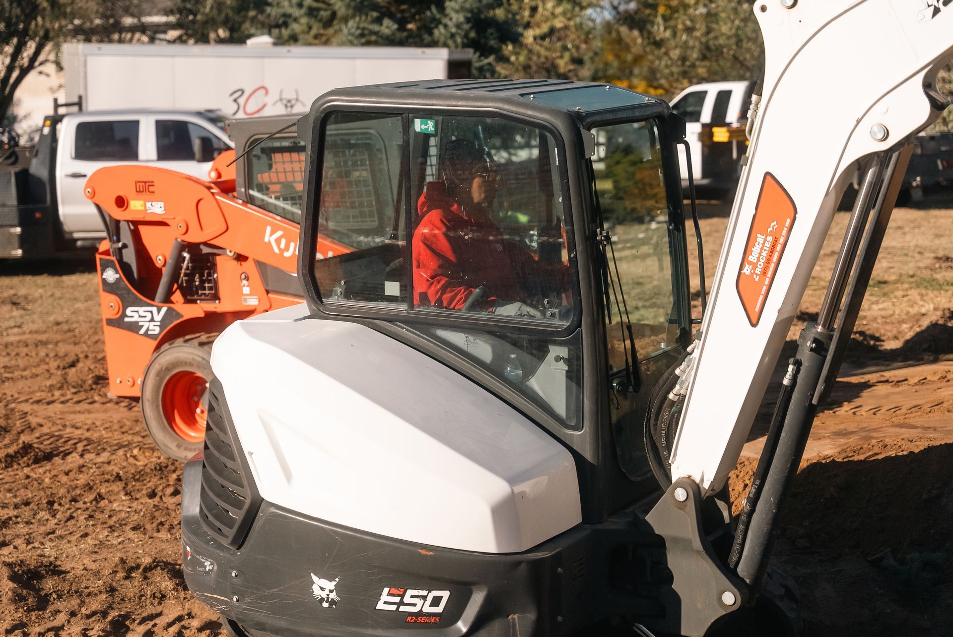 Person operating a Bobcat E50 excavator. Orange Kubota in background. Red shirt in the cab. Sunny outdoors.