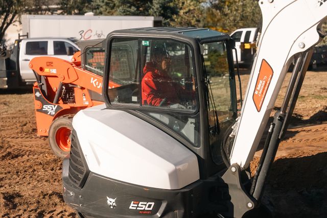 Person operating a Bobcat E50 excavator. Orange Kubota in background. Red shirt in the cab. Sunny outdoors.