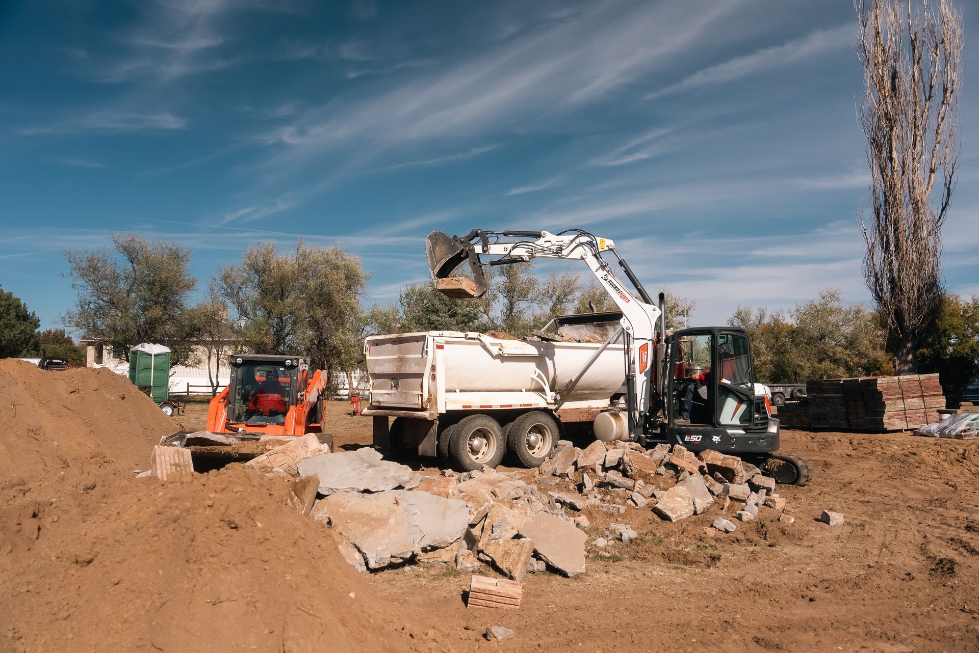 Construction site with a backhoe loading a dump truck. A small skid steer is nearby.