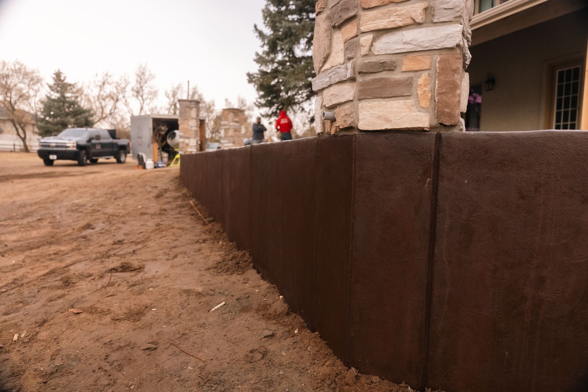 Brown concrete retaining wall in front of house; people and vehicles visible in the background.