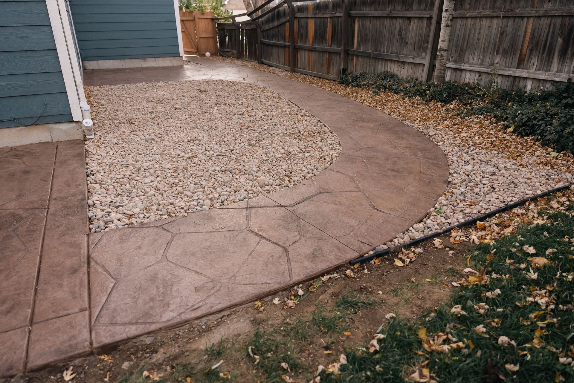 A concrete patio with a curved walkway, surrounded by gravel and grass, next to a blue building.