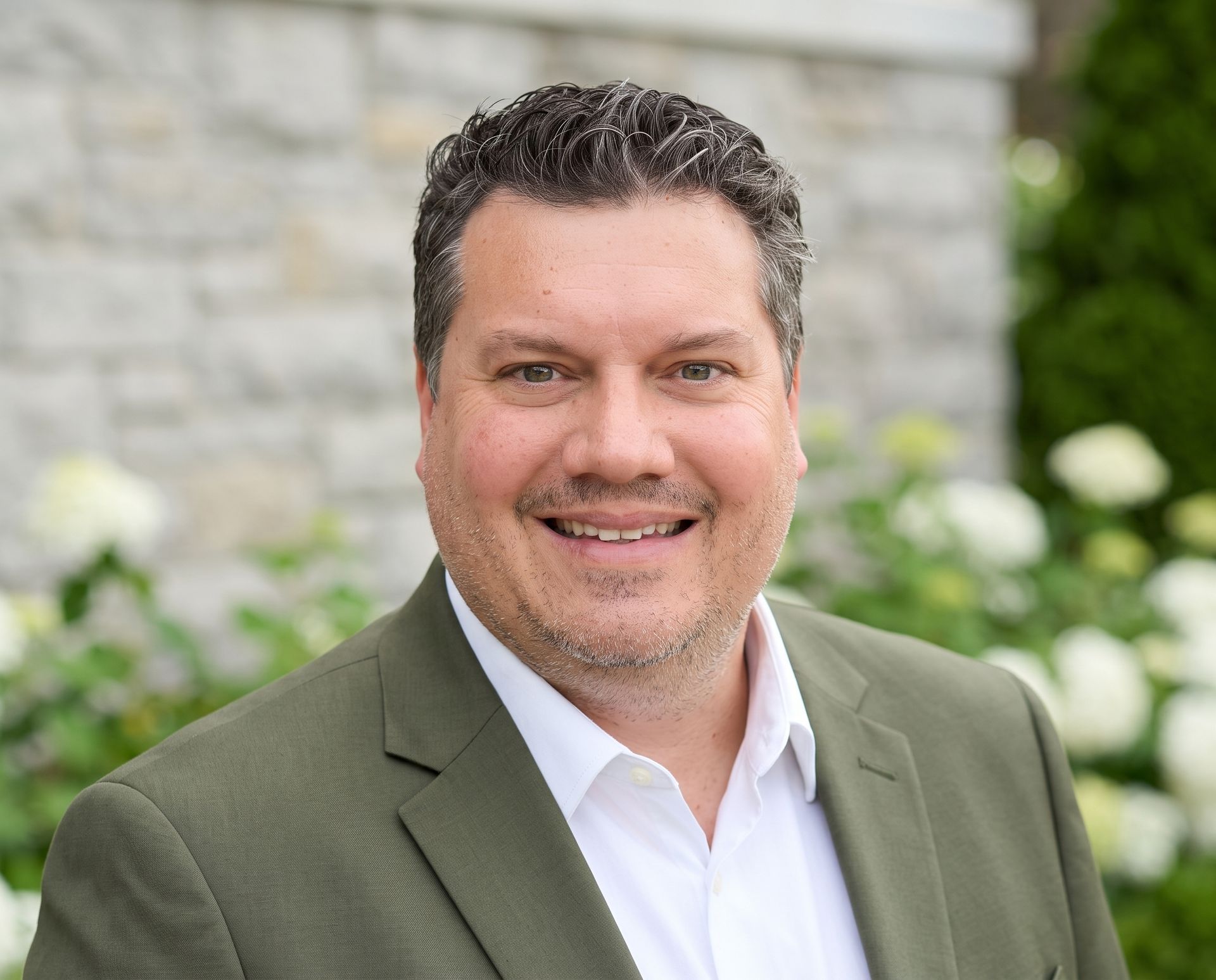 Smiling man in an olive blazer and white shirt, standing outdoors with blurred greenery and stone wall behind him