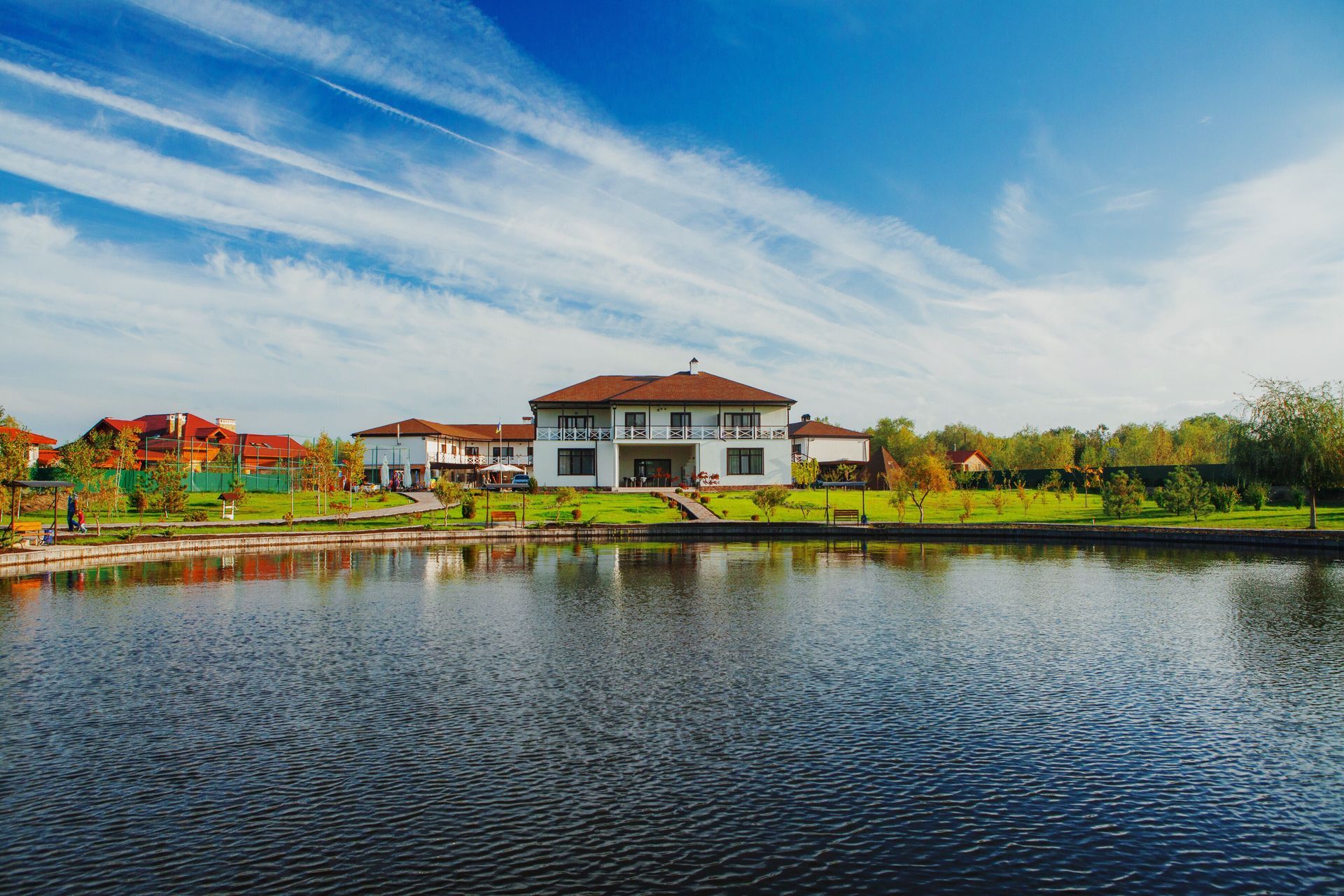 Large white house with brown roof reflected in a dark lake under a blue sky with clouds.
