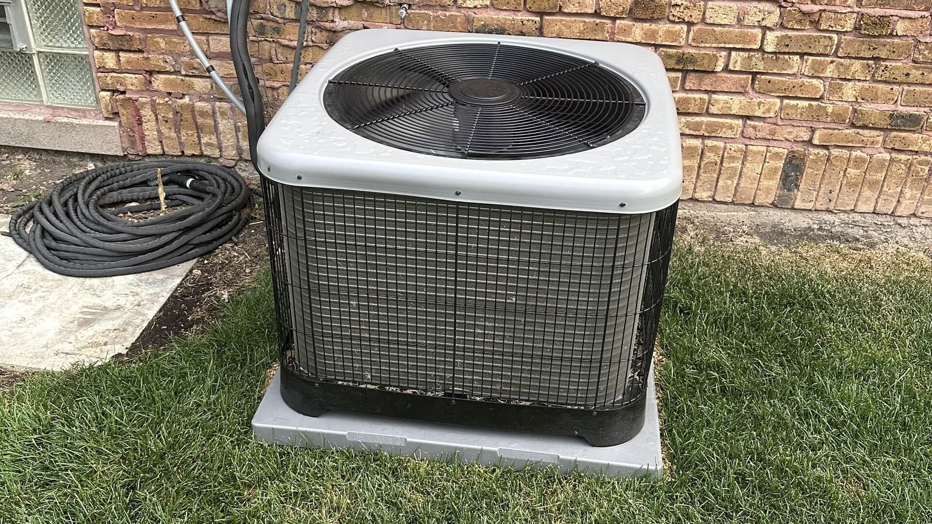 Outdoor air conditioner unit on grass beside a brick wall, with a coiled hose to the left.