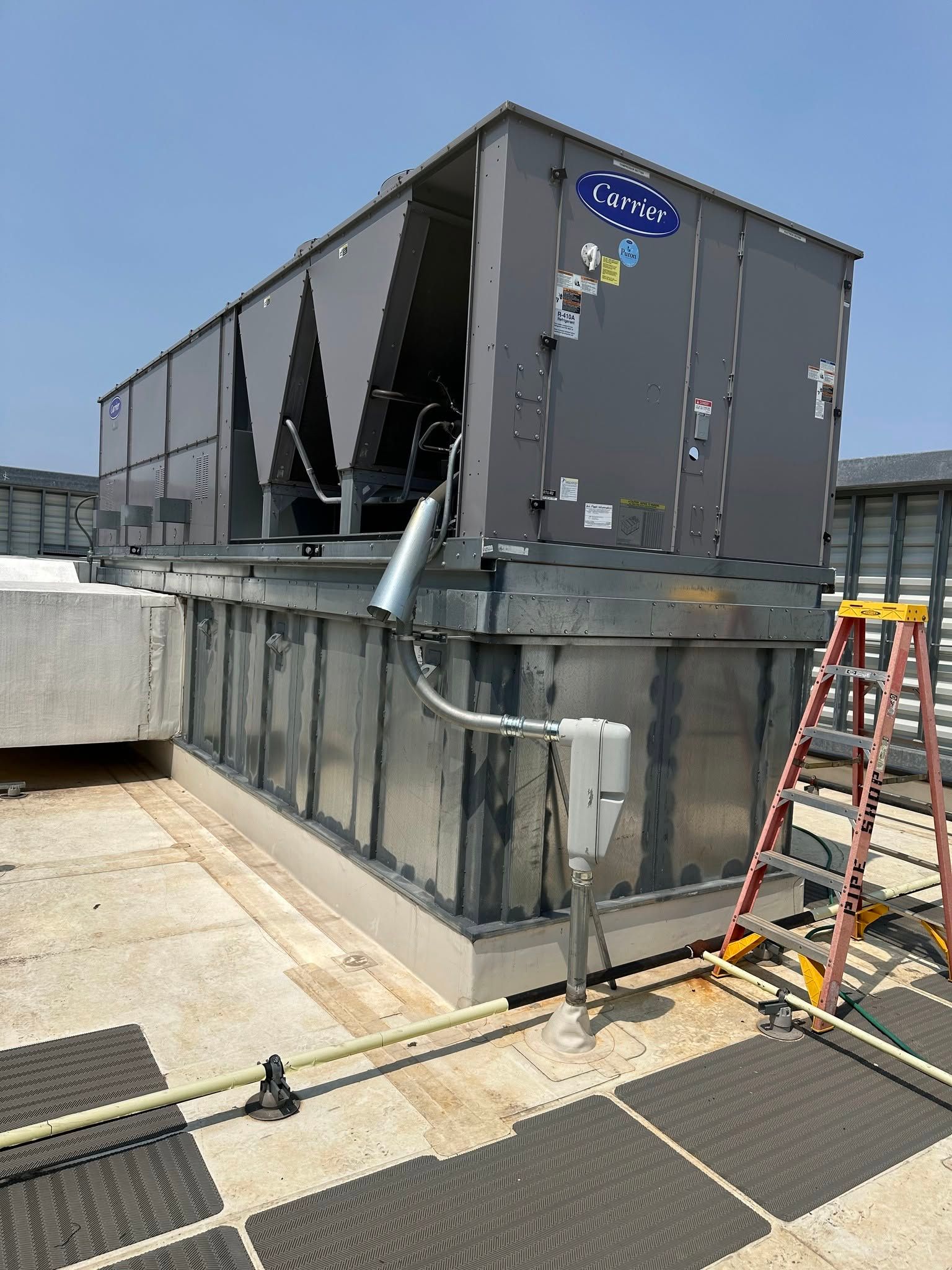 Rooftop HVAC unit beside concrete wall, with pipes and a red ladder on a sunny industrial rooftop.