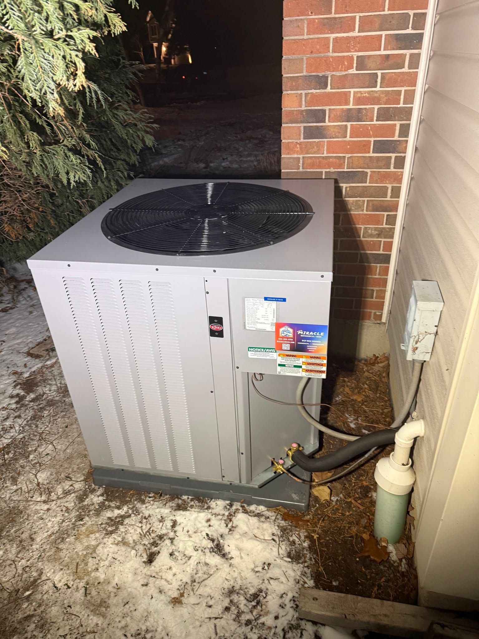 Outdoor HVAC unit beside a brick wall and doorway, with snow on the ground and exposed piping.