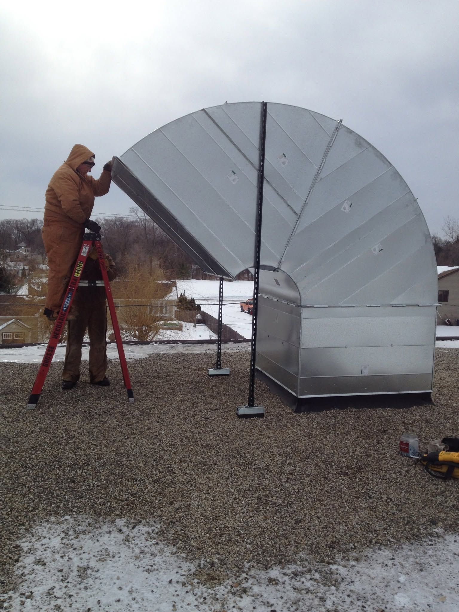 Worker on a ladder installing a large silver curved metal structure on a rooftop