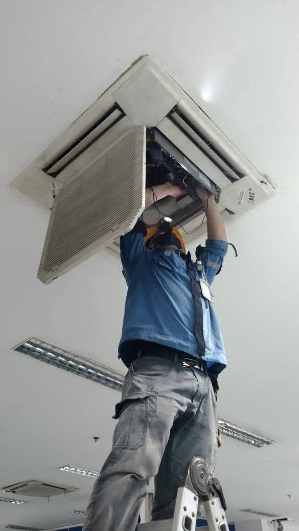 Worker on a ladder installing a ceiling air conditioner in a white room.