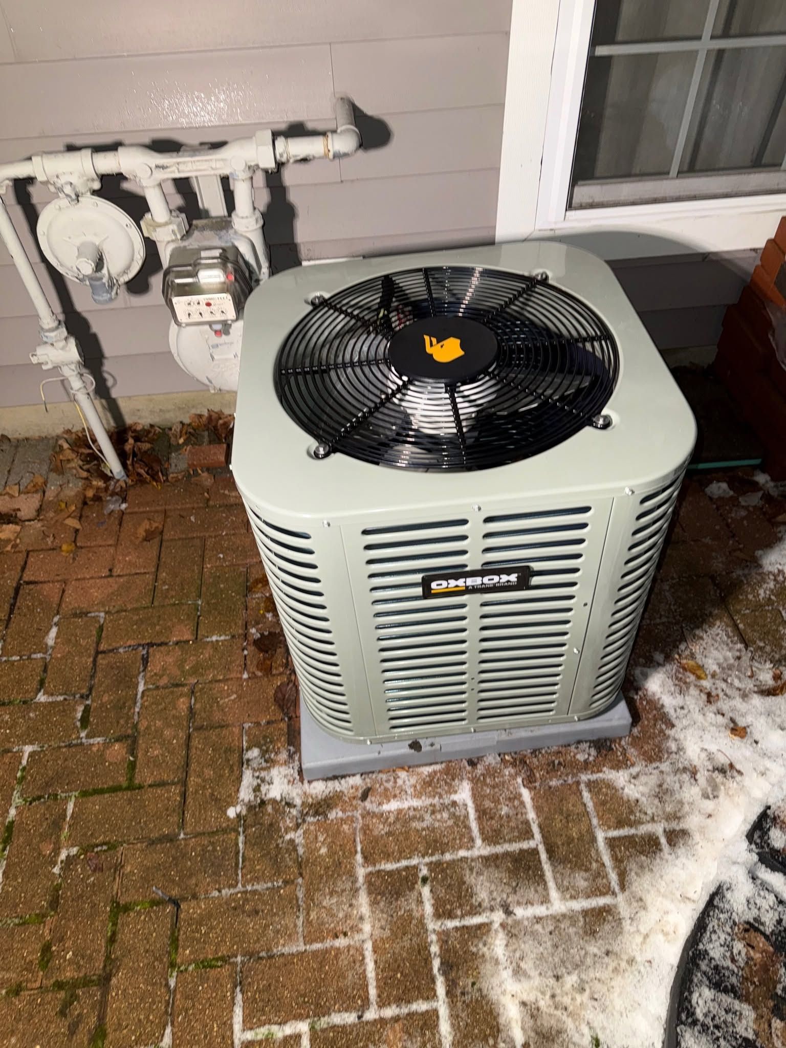 Outdoor HVAC condenser unit beside a brick wall, with snow and ice on the ground nearby.