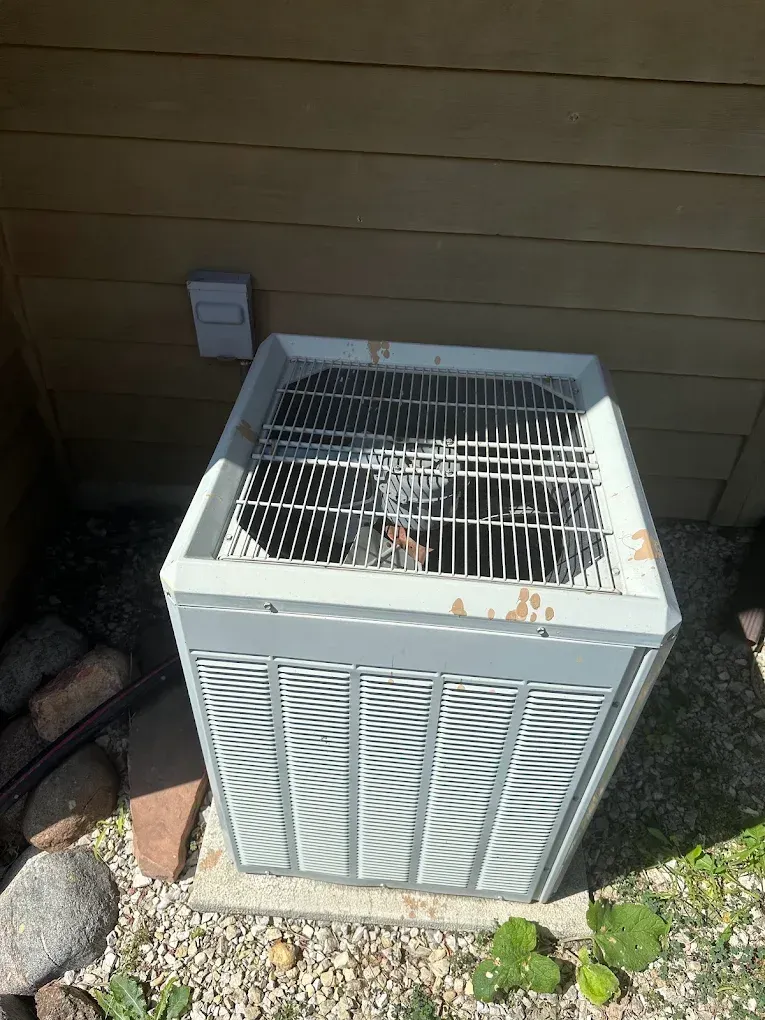 Outdoor air conditioner unit beside a house wall, with white grille and brown rust spots.