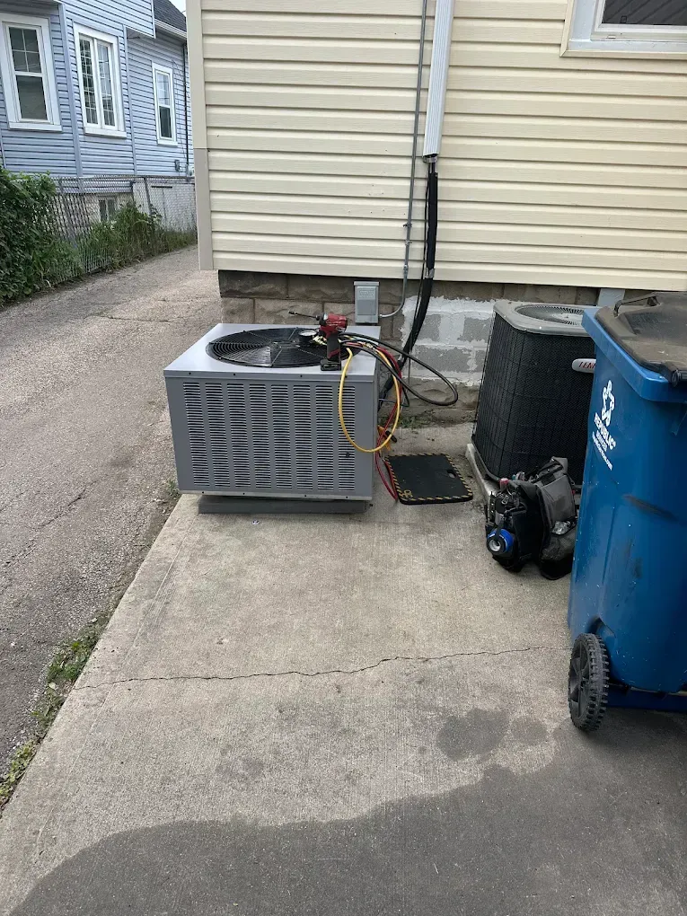 Outdoor AC unit beside a beige house, with a blue trash bin on a concrete driveway.
