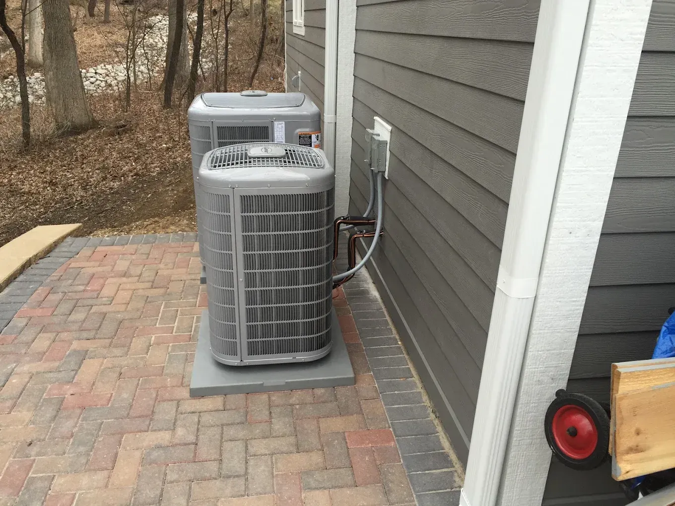 Outdoor air conditioning units beside a house on a brick patio, with a red blower on the right.