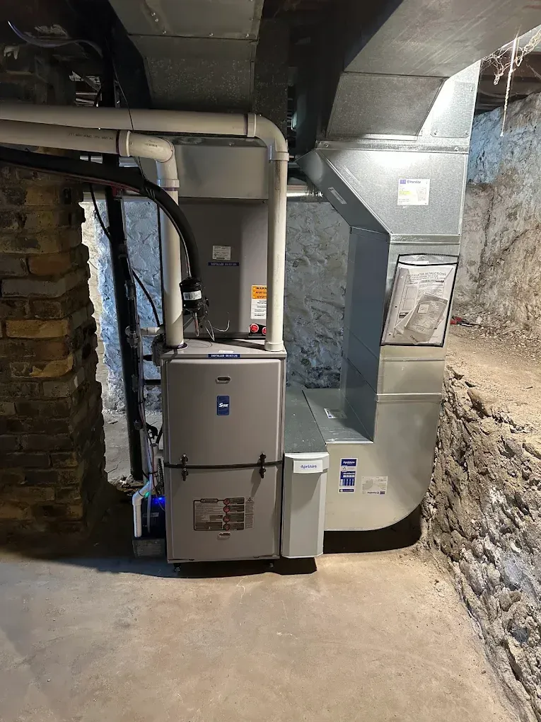 Large gray furnace and HVAC ductwork in a basement utility room with rough stone walls