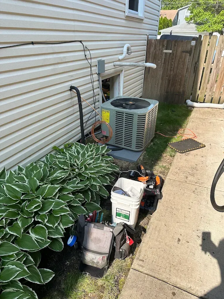 Outdoor HVAC unit and gardening supplies beside a house wall and hosta plants, next to a concrete walkway.
