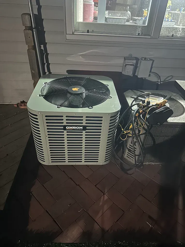 Outdoor air conditioner unit with exposed wiring beside a house window on a brick patio