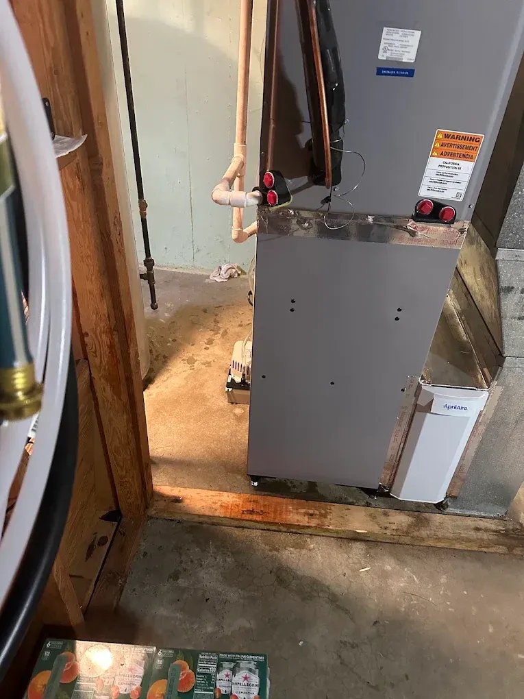 Utility room with a gray furnace, exposed pipes, and a concrete floor near a doorway.