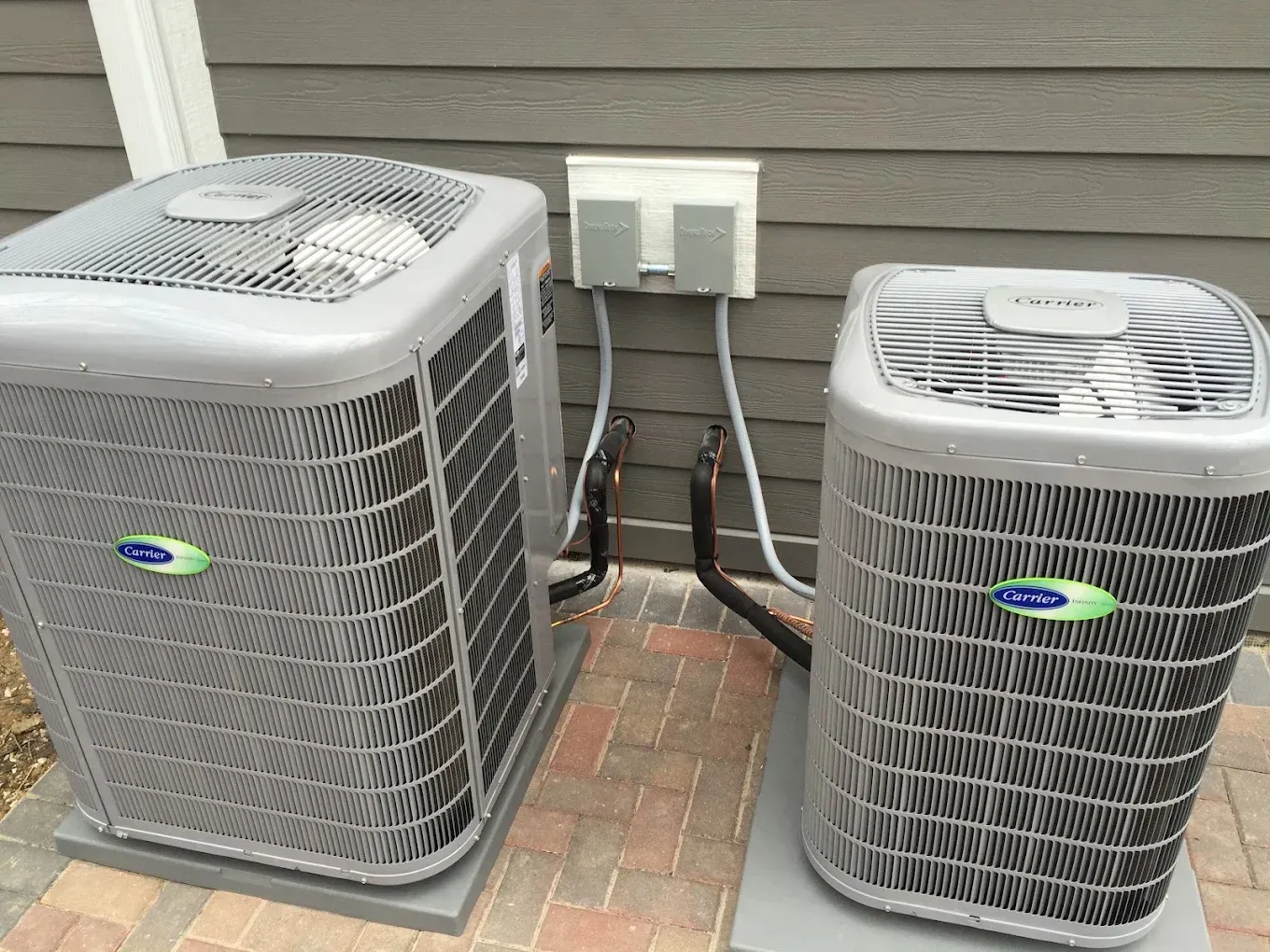 Two outdoor air conditioning units beside a house wall with exposed electrical conduits.