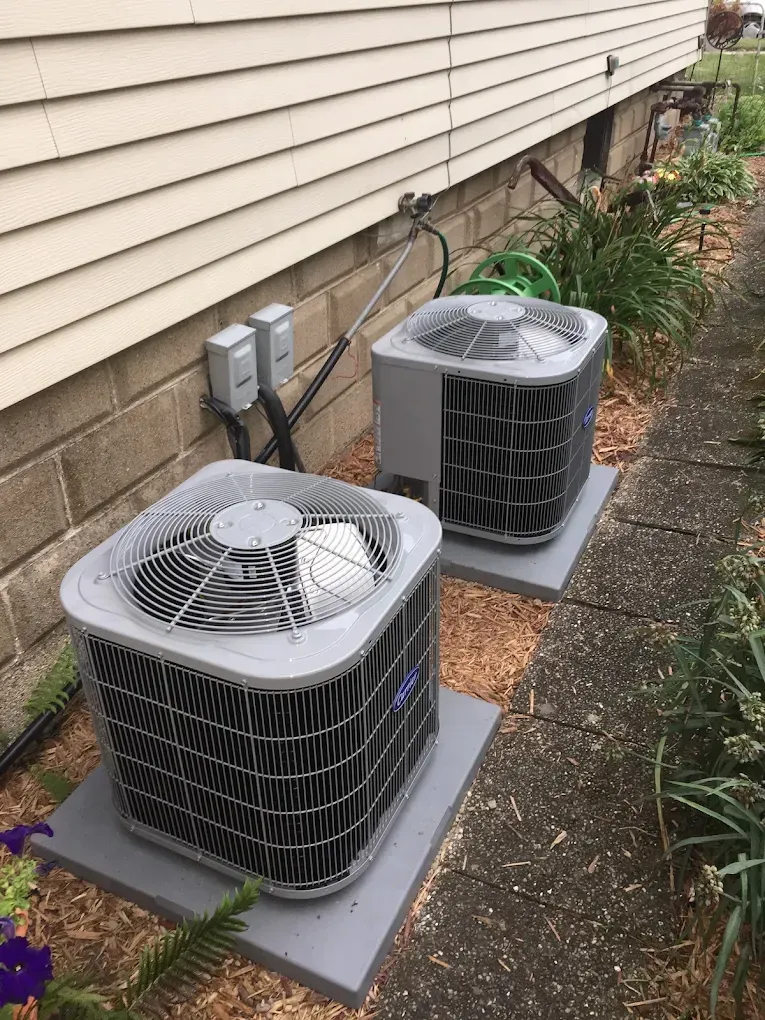 Two outdoor HVAC units beside a house with siding and brick foundation