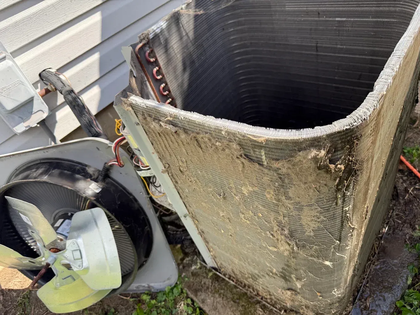 Damaged HVAC unit with exposed coils and detached fan beside a house exterior.