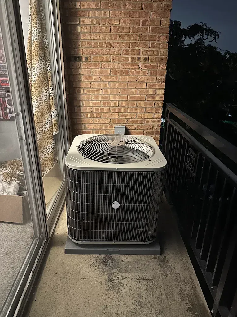 Outdoor HVAC unit on a narrow balcony beside a brick wall and sliding glass door at dusk
