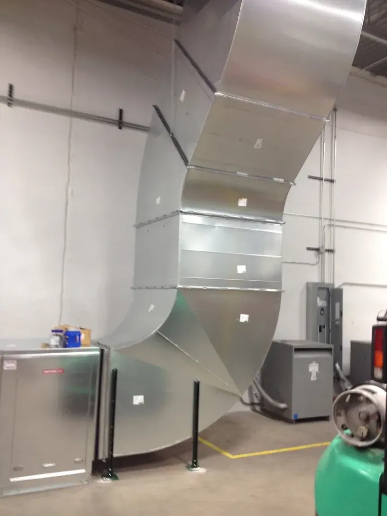 Large silver ventilation duct in an industrial room, with metal equipment cabinets and a green machine.