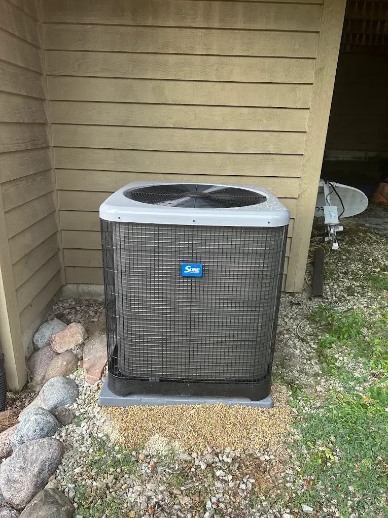 Outdoor air conditioner unit beside a beige house wall, with rocks and grass nearby.