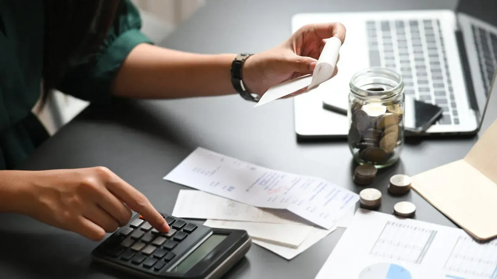 Person using a calculator and reviewing paperwork beside a laptop and jar of coins on a desk