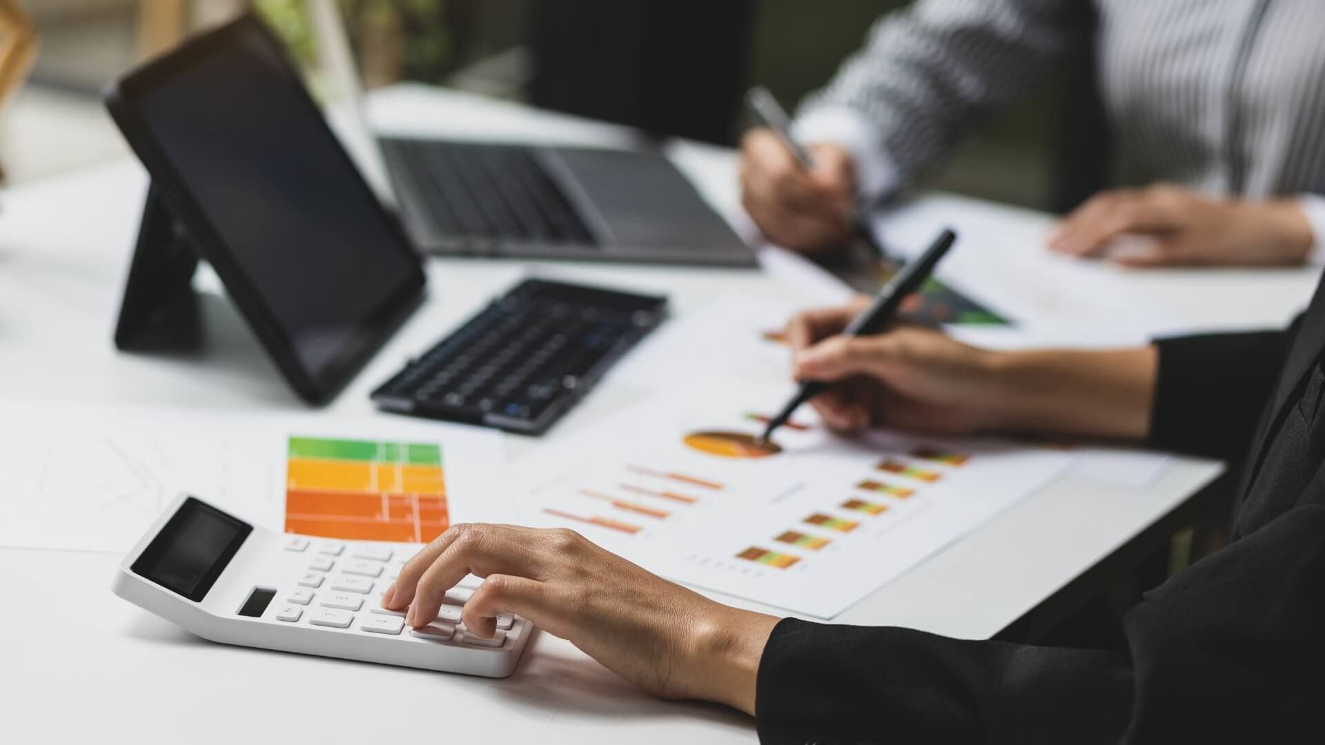 Hands reviewing colorful charts on a desk with laptops and a calculator during a business meeting
