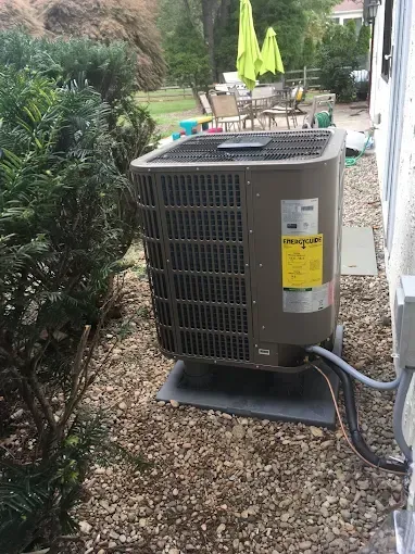 Outdoor air conditioner unit beside a house, with shrubs and patio furniture in the background