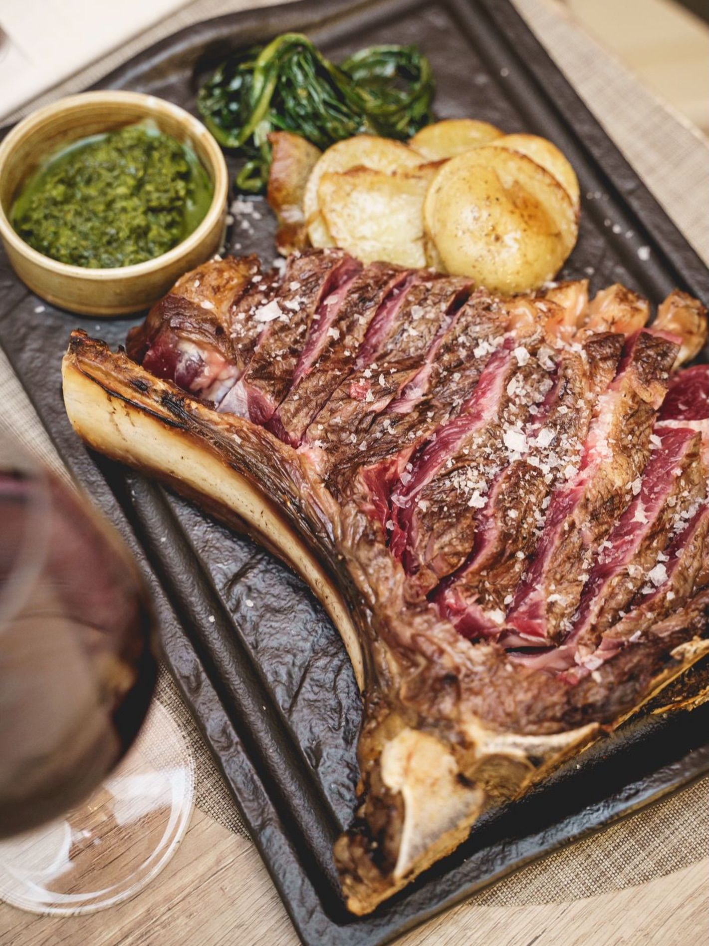 Bistecca tomahawk alla griglia con patate, verdure e salsa su un piatto nero.