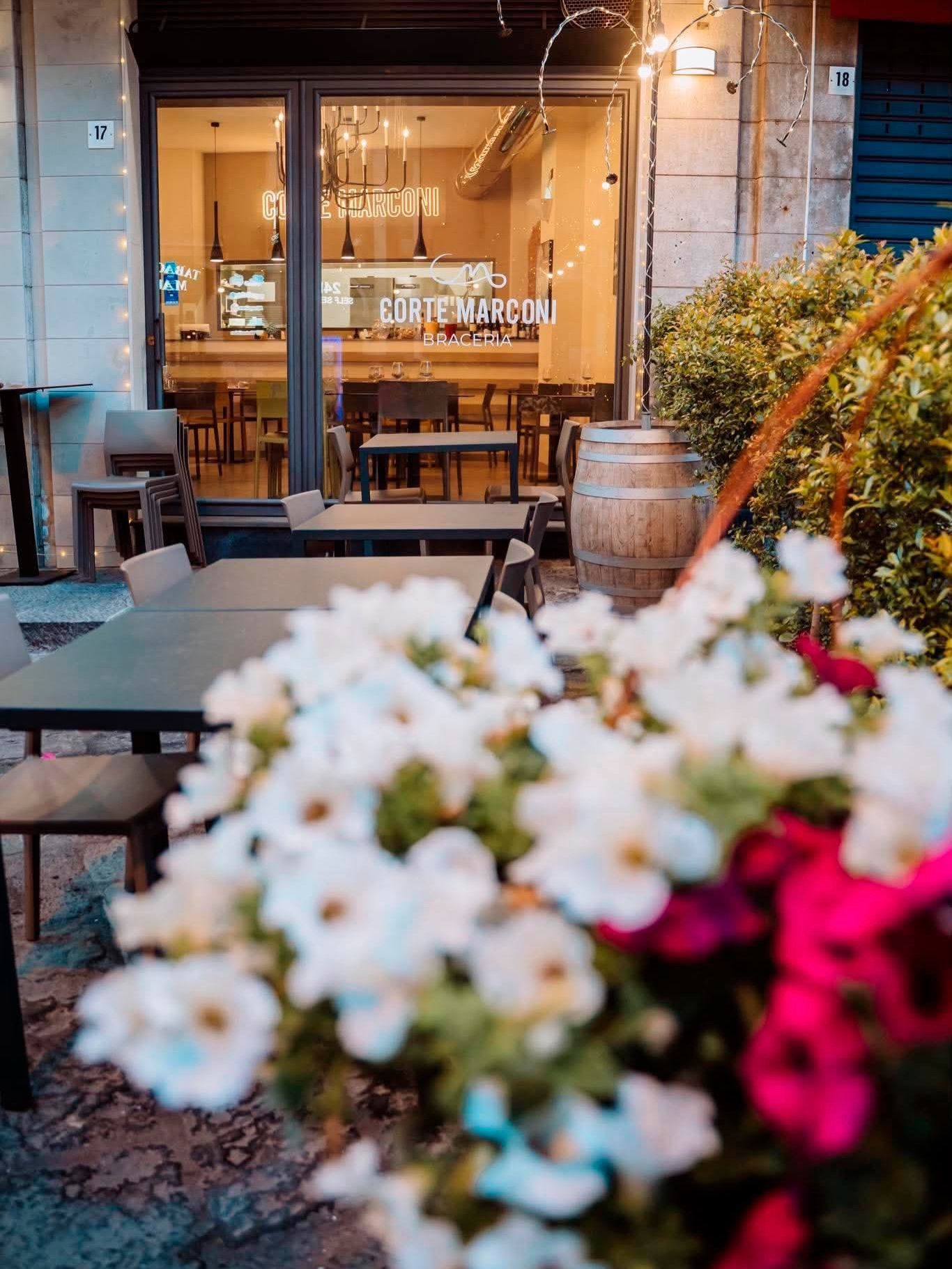 Accogliente ingresso di un caffè con luci decorative, tavolini in legno e fiori bianchi e rosa sfocati in primo piano.