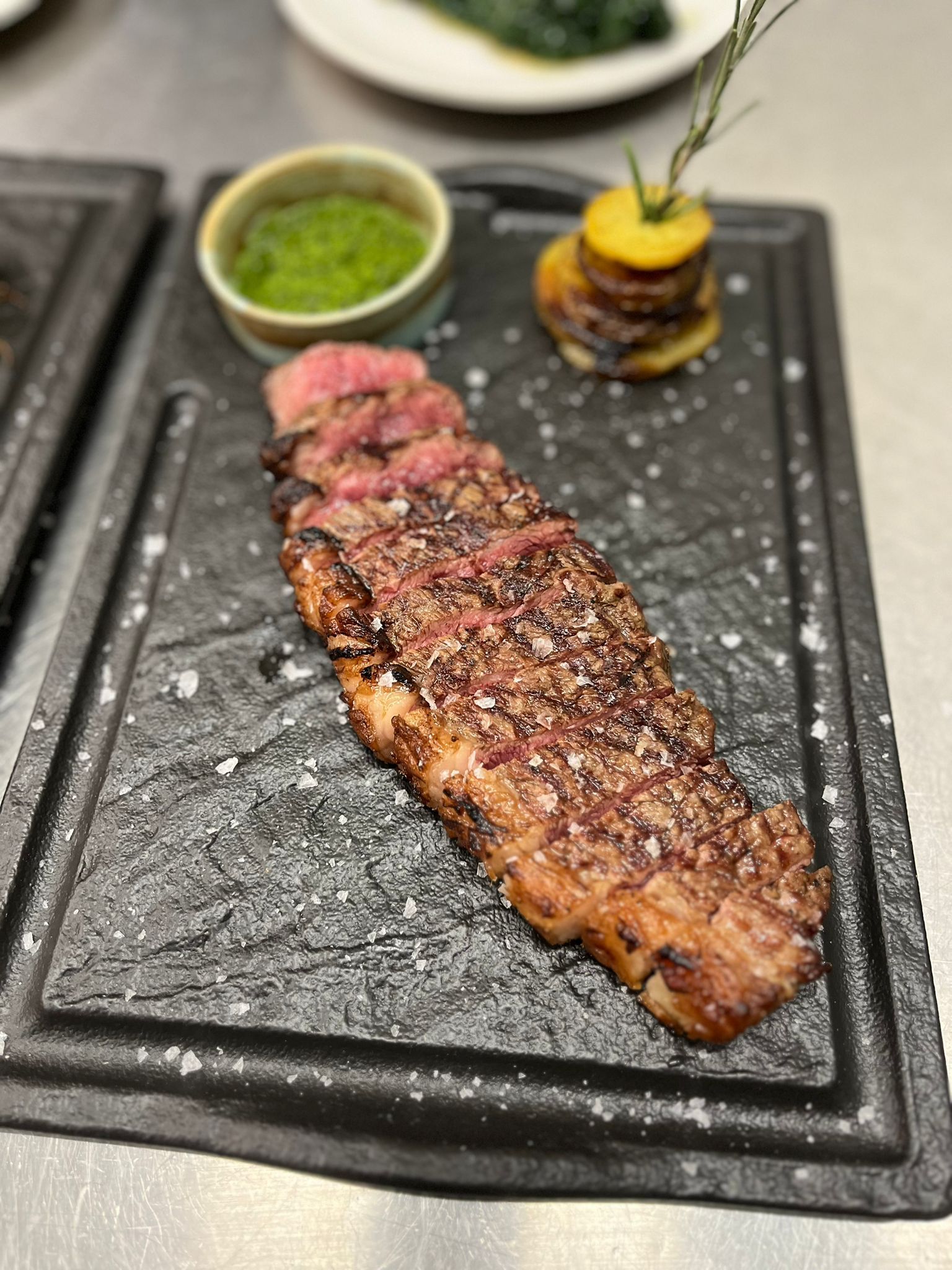 Bistecca alla griglia su un piatto di ardesia nera con salsa verde e una piccola ciotola di guarnizione.
