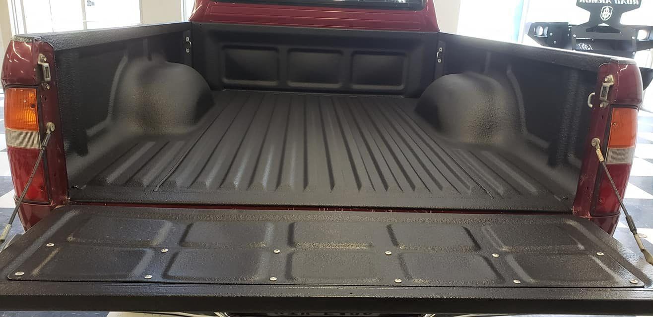 The open bed of a maroon pickup truck, featuring a black spray-in bed liner and a textured tailgate.